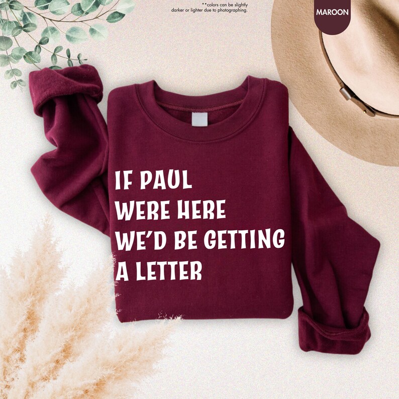 May include: Maroon sweatshirt with the text "IF PAUL WERE HERE WE'D BE GETTING A LETTER" in white. The sleeves are rolled up. The word "MAROON" is in a circle in the upper right corner. The background has a hat and some greenery.