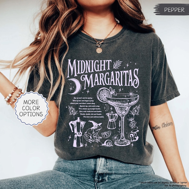 May include: Dark gray t-shirt with the words "Midnight Margaritas" in a decorative font, along with a margarita glass illustration and other botanical elements. The shirt also has the text "More Color Options".