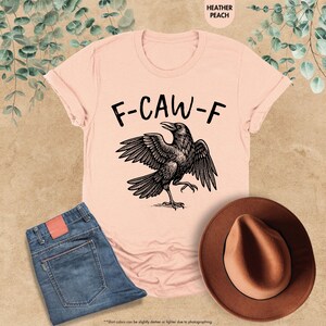 May include: A heather peach t-shirt with the text "F-CAW-F" above a black raven illustration. The image also includes a pair of blue jeans and a brown hat. The shirt's color may vary slightly due to photography.