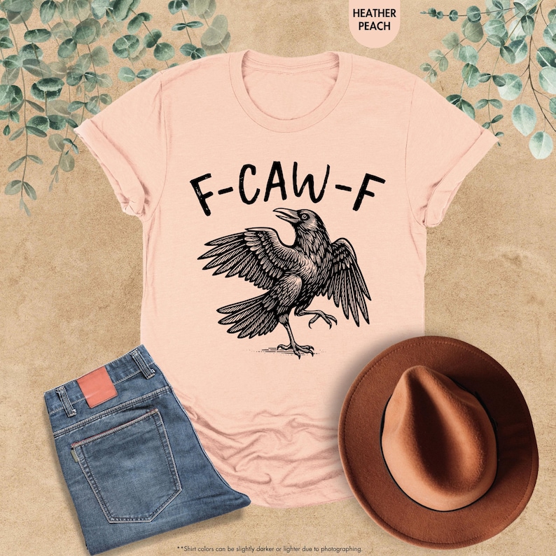 May include: A heather peach t-shirt with the text "F-CAW-F" above a black raven illustration. The image also includes a pair of blue jeans and a brown hat. The shirt's color may vary slightly due to photography.