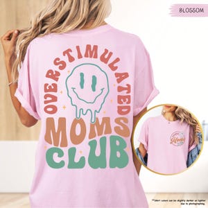 K&ouml;nnte beinhalten: Hellrosa T-Shirt mit dem Schriftzug "OVERSTIMULATED MOMS CLUB" in Retro-Schrift. Das Design zeigt ein schmelzendes Smiley-Gesicht. Das Shirt hat kurze &Auml;rmel und einen Rundhalsausschnitt.