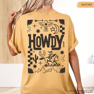 May include: A mustard yellow t-shirt with a black graphic print of a cowboy riding a horse, a horseshoe, a cactus, stars, and the word "HOWDY".
