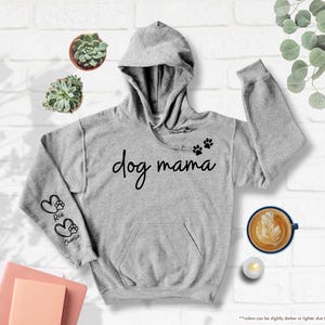 May include: A light grey hoodie with the text "dog mama" in black script. The hoodie features paw prints and the names "Rex" and "Charlie" on the sleeves. The hoodie has a hood and a front pocket.