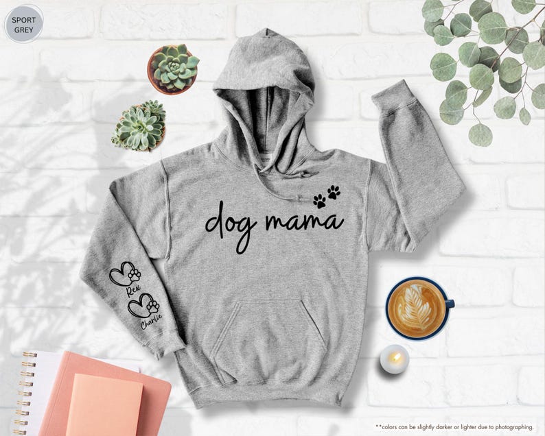 May include: A light grey hoodie with the text "dog mama" in black script. The hoodie features paw prints and the names "Rex" and "Charlie" on the sleeves. The hoodie has a hood and a front pocket.