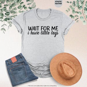 Pode incluir: Uma camiseta cinza claro com o texto "WAIT FOR ME i have little legs" em preto. A camiseta &eacute; combinada com jeans azuis e um chap&eacute;u marrom. As palavras "ATHLETIC HEATHER" est&atilde;o no topo.