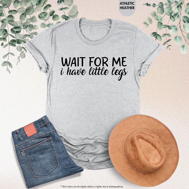 Pode incluir: Uma camiseta cinza claro com o texto "WAIT FOR ME i have little legs" em preto. A camiseta &eacute; combinada com jeans azuis e um chap&eacute;u marrom. As palavras "ATHLETIC HEATHER" est&atilde;o no topo.