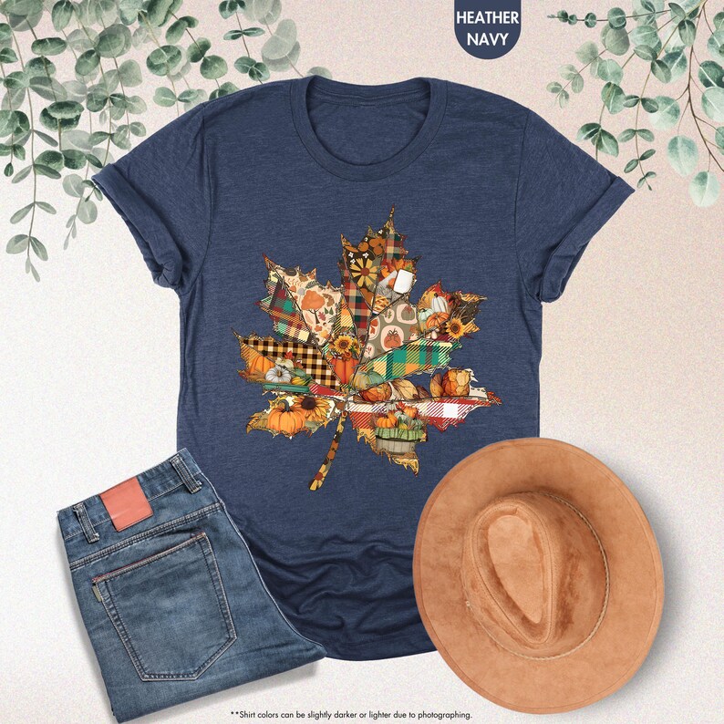 Puede incluir: Una camiseta de manga corta en azul marino jaspeado con un gr&aacute;fico de hoja de arce compuesto por varios patrones oto&ntilde;ales, incluyendo calabazas, girasoles y cuadros, presentada junto a unos vaqueros azules y un sombrero de fieltro color canela.