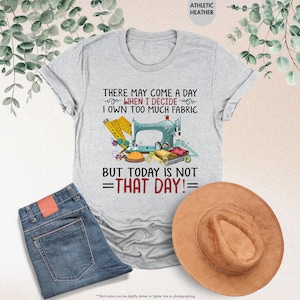 May include: A gray t-shirt with a sewing machine and fabric design. The text reads "There may come a day when I decide I own too much fabric but today is not that day!"
