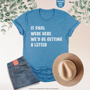 May include: A teal t-shirt with the text "IF PAUL WERE HERE WE'D BE GETTING A LETTER" in white, paired with blue jeans and a tan fedora hat. The shirt is folded at the sleeves. The image includes a color swatch that says "HEATHER DEEP TEAL".