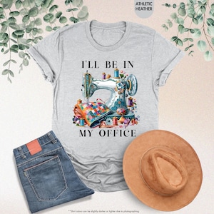 K&ouml;nnte beinhalten: Ein graues T-Shirt mit dem Text "I'll be in my office" und einer bunten Illustration einer N&auml;hmaschine mit Garn und Stoff.