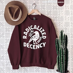 May include: Maroon sweatshirt with the text "RADICALIZED BY BASIC DECENCY" in a white, circular design. The design includes a dove holding a sprig. A brown hat with a white band hangs to the left.