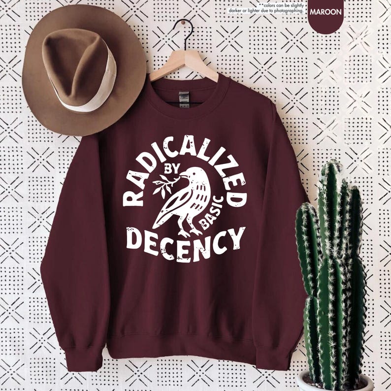 May include: Maroon sweatshirt with the text "RADICALIZED BY BASIC DECENCY" in a white, circular design. The design includes a dove holding a sprig. A brown hat with a white band hangs to the left.