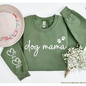 May include: A military green sweatshirt with the words "dog mama" in white script, and two white paw prints. The left sleeve has a heart and paw print design with the names "Rex" and "Charlie". A beige hat with a floral band is on the left. A small bouquet of white flowers is on the right.