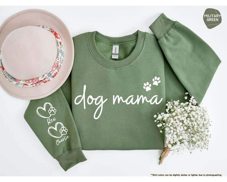May include: A military green sweatshirt with the words "dog mama" in white script, and two white paw prints. The left sleeve has a heart and paw print design with the names "Rex" and "Charlie". A beige hat with a floral band is on the left. A small bouquet of white flowers is on the right.