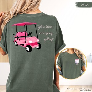 May include: Moss green t-shirt with a pink golf cart graphic and the text "get in losers we're going golfing." The shirt also has a small graphic of a golf ball with a pink bow on the front. The shirt is made of a soft material.