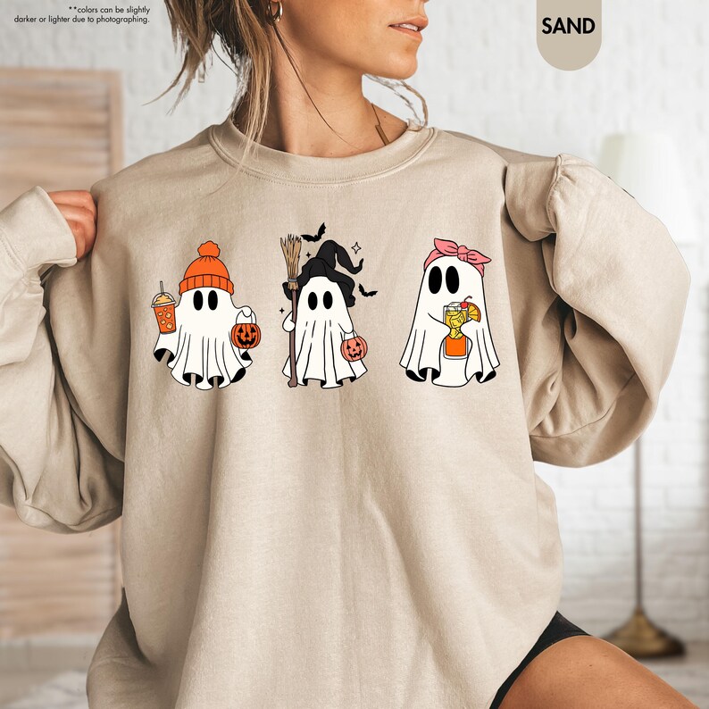 May include: A sand-coloured crewneck sweatshirt featuring a graphic print of three white ghosts. The first ghost wears an orange beanie and holds a drink and pumpkin bucket. The second wears a witch hat and holds a broom. The third wears a pink headband.