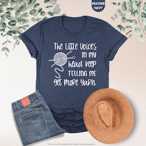 May include: A navy blue t-shirt with white text that reads "The Little Voices In My Head Keep Telling Me Get More Yarn". The text is accompanied by a graphic of a ball of yarn with knitting needles.