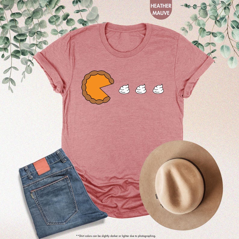 May include: A heather mauve t-shirt with a graphic of a pumpkin pie slice with a bite taken out of it and three dollops of whipped cream. The shirt is displayed on a wooden surface with a pair of blue jeans and a brown hat.