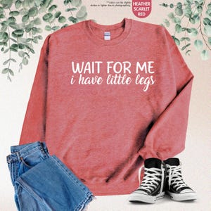 Pode incluir: Camisola vermelha escarlate mesclada com o texto "WAIT FOR ME i have little legs" em branco. A imagem tamb&eacute;m mostra cal&ccedil;as de ganga azuis e t&eacute;nis pretos e brancos.