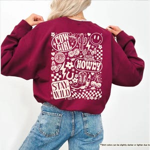 K&ouml;nnte beinhalten: Ein weinrotes Sweatshirt mit einem wei&szlig;en Grafikdesign auf dem R&uuml;cken. Das Design enth&auml;lt die W&ouml;rter "Cow Girl", "Howdy" und "Stay Wild" sowie Bilder von Kaktus, Stiefeln, Schmetterlingen und einem Smiley.