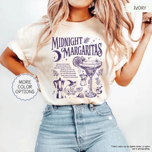 May include: Ivory-colored t-shirt with the text "MIDNIGHT MARGARITAS" in dark blue. The design includes a cocktail glass, a crescent moon, and other illustrations. The shirt also has the words "MORE COLOR OPTIONS" on it.