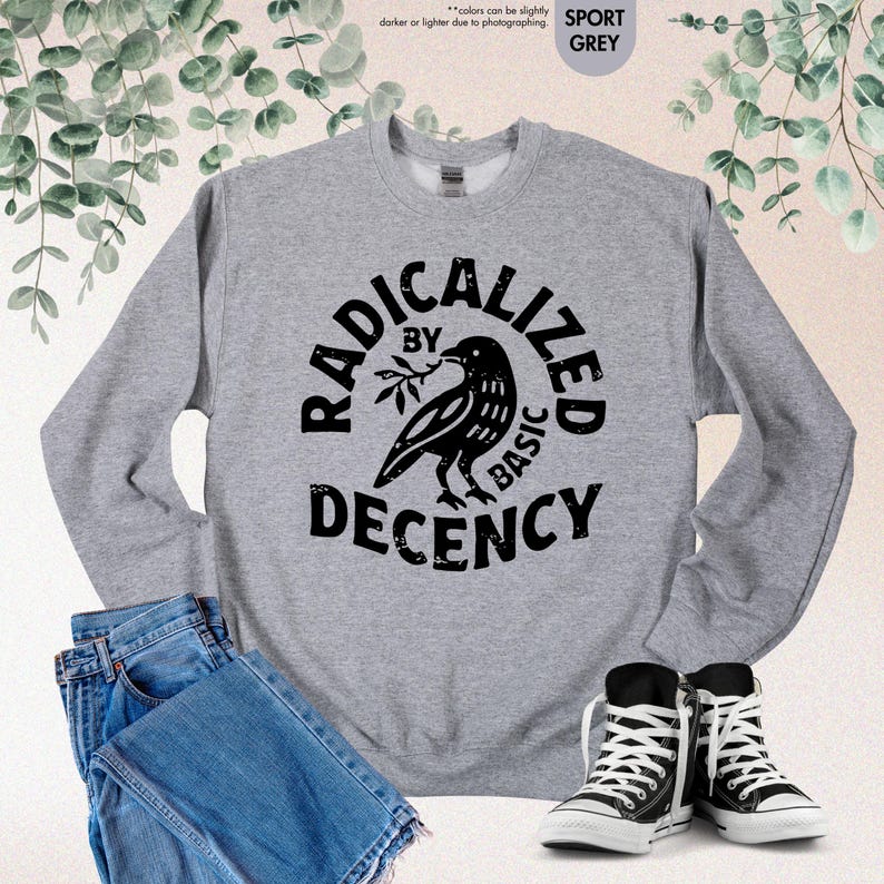 May include: Sport grey sweatshirt with the text "RADICALIZED BY BASIC DECENCY" in a circular design with a black bird graphic. The sweatshirt is paired with blue jeans and black and white sneakers.