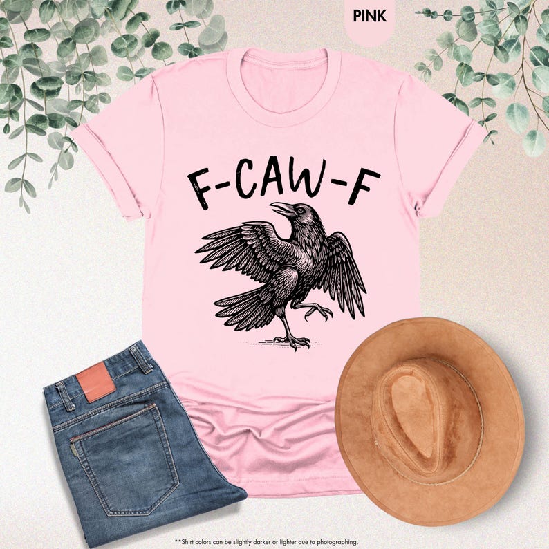 May include: Pink t-shirt with the text "F-CAW-F" above a black raven illustration. The image also includes a pair of blue jeans and a brown hat. The shirt's color is pink.