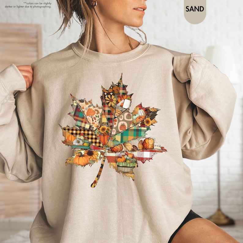 Puede incluir: Una sudadera de cuello redondo color arena con un gran gr&aacute;fico central de una hoja de arce. La hoja est&aacute; compuesta por varios patrones de patchwork, incluyendo cuadros, vichy y estampados florales en naranja, amarillo, verde y marr&oacute;n.
