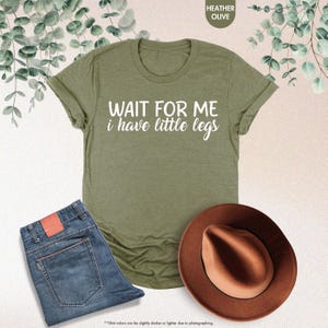 Pode incluir: T-shirt verde-oliva com o texto "WAIT FOR ME i have little legs" em branco. A imagem tamb&eacute;m mostra um par de jeans azuis e um chap&eacute;u fedora castanho.