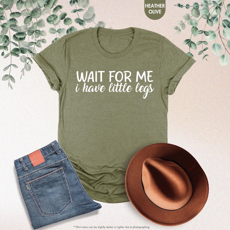 Pode incluir: T-shirt verde-oliva com o texto "WAIT FOR ME i have little legs" em branco. A imagem tamb&eacute;m mostra um par de jeans azuis e um chap&eacute;u fedora castanho.