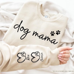 May include: A sand-colored sweatshirt with the words "dog mama" in a black script font. Two black paw prints are below the word "mama". The sleeve features paw prints inside heart shapes with the names "Rex" and "Charlie".