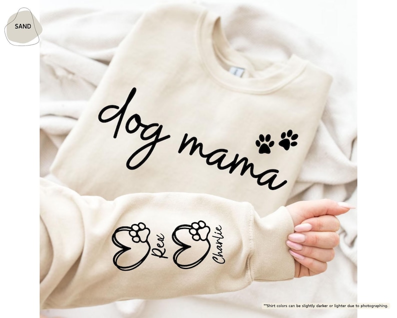 May include: A sand-colored sweatshirt with the words "dog mama" in a black script font. Two black paw prints are below the word "mama". The sleeve features paw prints inside heart shapes with the names "Rex" and "Charlie".