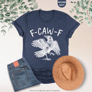 May include: Heather navy t-shirt with a white graphic of a crow and the text "F-CAW-F". The image also shows a pair of blue jeans and a brown hat. The shirt is made of a soft material.