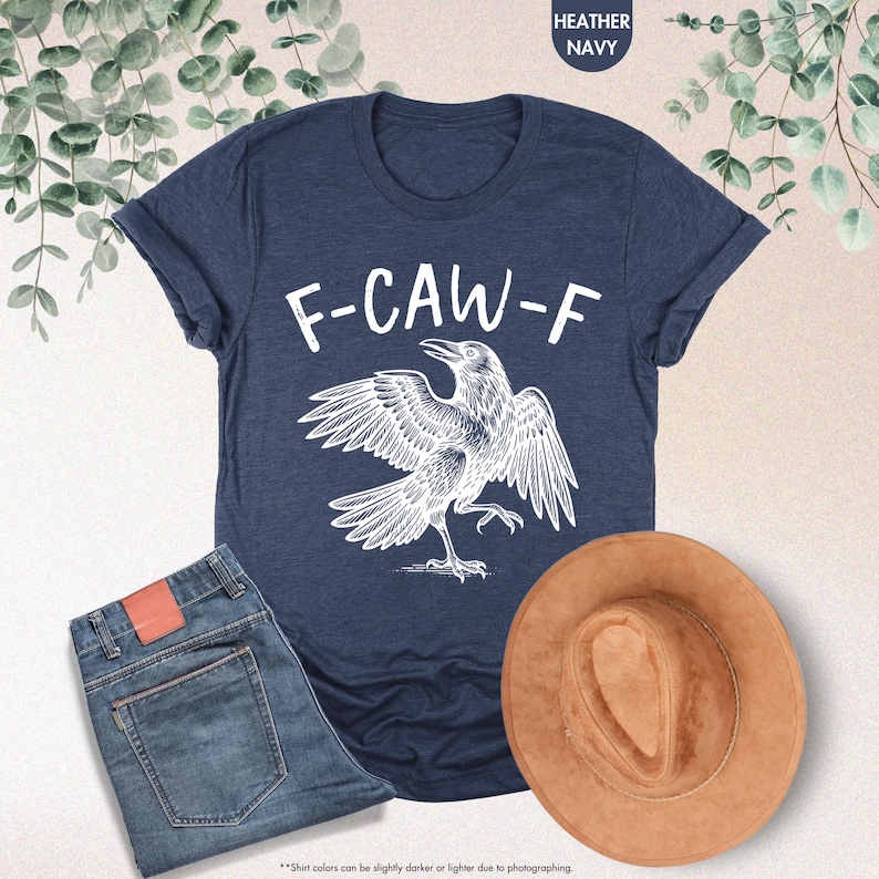 May include: Heather navy t-shirt with a white graphic of a crow and the text "F-CAW-F". The image also shows a pair of blue jeans and a brown hat. The shirt is made of a soft material.