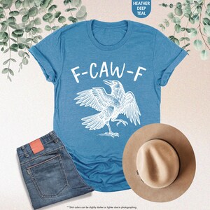 May include: A teal t-shirt with the text "F-CAW-F" and a white raven graphic. The shirt is paired with blue jeans and a tan fedora hat. The image includes the text "HEATHER DEEP TEAL".