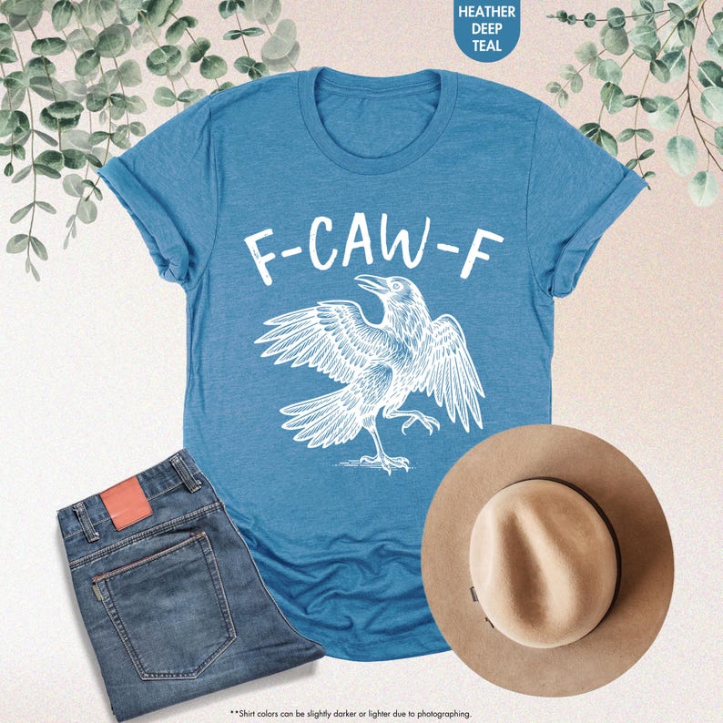 May include: A teal t-shirt with the text "F-CAW-F" and a white raven graphic. The shirt is paired with blue jeans and a tan fedora hat. The image includes the text "HEATHER DEEP TEAL".