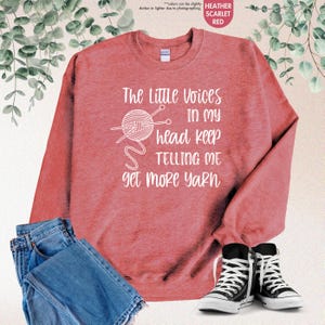 May include: A heather scarlet red sweatshirt with the text "The Little Voices In My Head Keep Telling Me Get More Yarn" in white. A white outline of a ball of yarn with knitting needles is above the text.