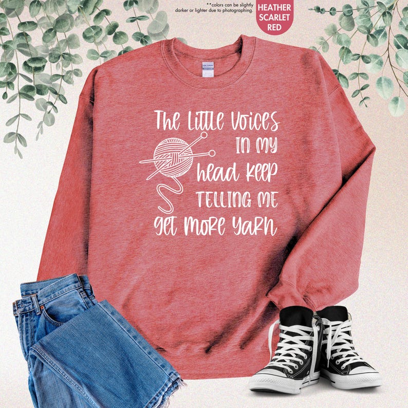 May include: A heather scarlet red sweatshirt with the text "The Little Voices In My Head Keep Telling Me Get More Yarn" in white. A white outline of a ball of yarn with knitting needles is above the text.