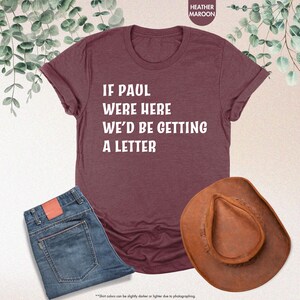 May include: Heather maroon t-shirt with the text "IF PAUL WERE HERE WE'D BE GETTING A LETTER". The image also includes a pair of blue jeans and a brown cowboy hat. The shirt is a casual wear item.