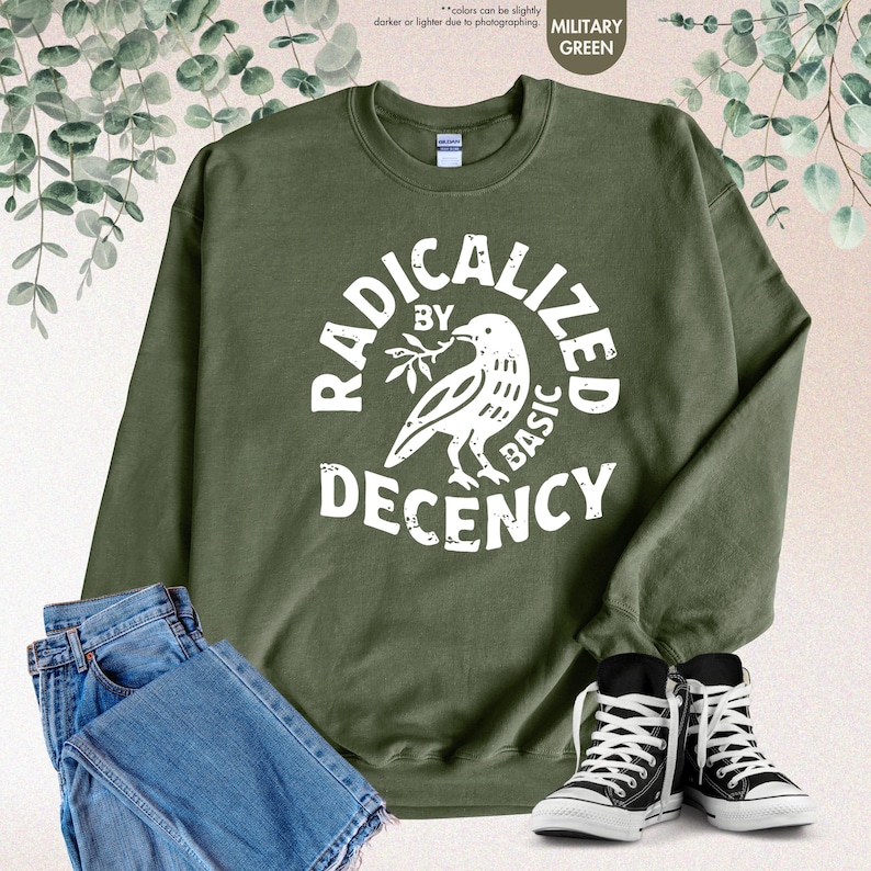 May include: Military green sweatshirt with the text "RADICALIZED BY BASIC DECENCY" in white. The design includes a dove holding a branch. The sweatshirt is paired with blue jeans and black and white sneakers.