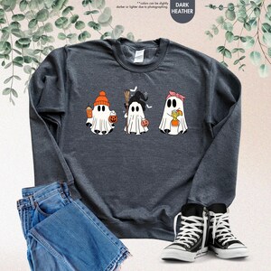 May include: A dark heather grey crewneck sweatshirt featuring a graphic of three white ghosts. The first ghost wears an orange beanie and holds a drink and pumpkin bucket. The second wears a witch hat and holds a broom. The third wears a pink headband.