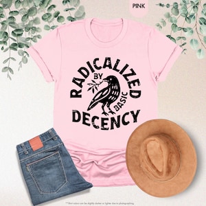 May include: A pink t-shirt with the words "RADICALIZED BY BASIC DECENCY" in black, featuring a raven illustration. The shirt is paired with blue jeans and a brown hat. The text "PINK" is in the top right corner.