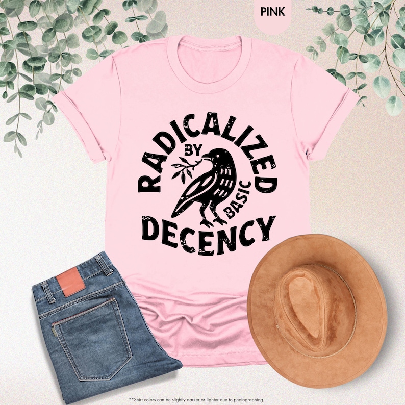 May include: A pink t-shirt with the words "RADICALIZED BY BASIC DECENCY" in black, featuring a raven illustration. The shirt is paired with blue jeans and a brown hat. The text "PINK" is in the top right corner.