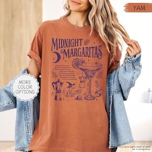 May include: A yam-colored t-shirt with the words "Midnight Margaritas" in dark blue. The graphic includes a cocktail, moon, frog, and other illustrations. The shirt has rolled-up sleeves.