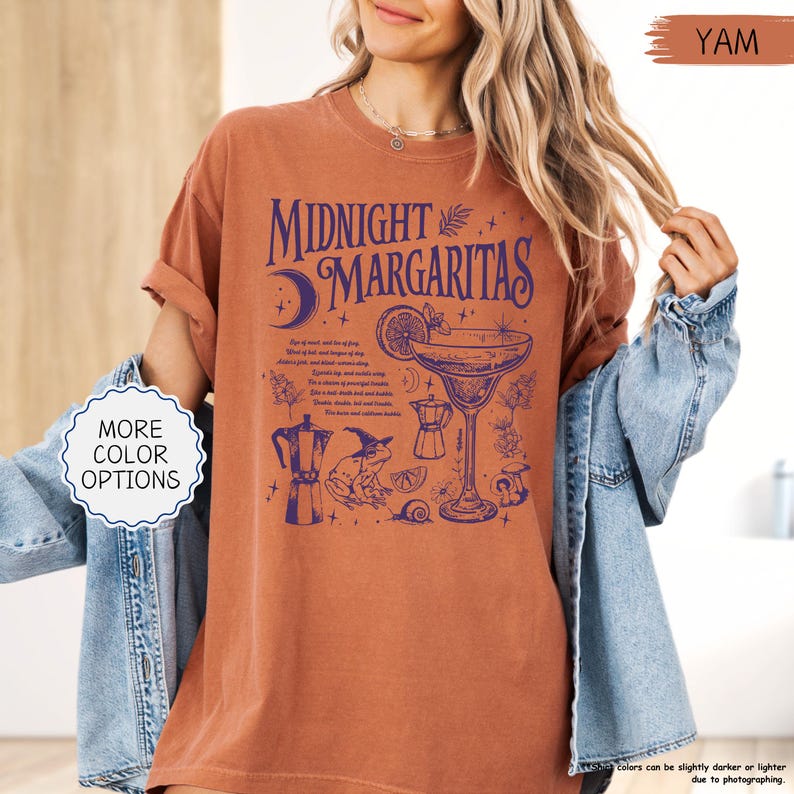 May include: A yam-colored t-shirt with the words "Midnight Margaritas" in dark blue. The graphic includes a cocktail, moon, frog, and other illustrations. The shirt has rolled-up sleeves.