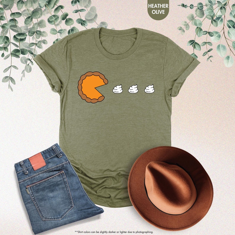 May include: A green t-shirt with a graphic of a pumpkin pie with a bite taken out of it and three dollops of whipped cream. The text "HEATHER OLIVE" is at the top of the image.