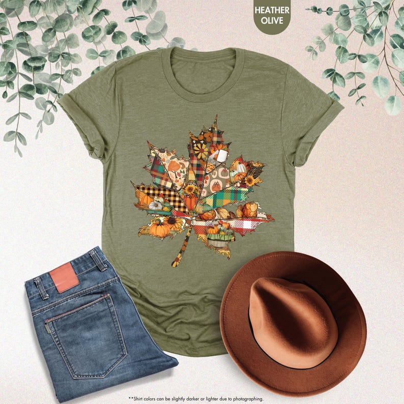Puede incluir: Una camiseta color oliva jaspeado con un gr&aacute;fico grande de hoja de arce compuesto por varios segmentos estampados que incluyen calabazas, girasoles, cuadros y flores. La prenda se muestra junto a unos vaqueros azules y un sombrero fedora marr&oacute;n.