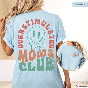 K&ouml;nnte beinhalten: Hellblaues T-Shirt mit dem Text "OVERSTIMULATED MOMS CLUB" in Orange, Gr&uuml;n und Rot. Das Design beinhaltet ein schmelzendes Smiley-Gesicht. Das Shirt hat die Farbe Chambray.