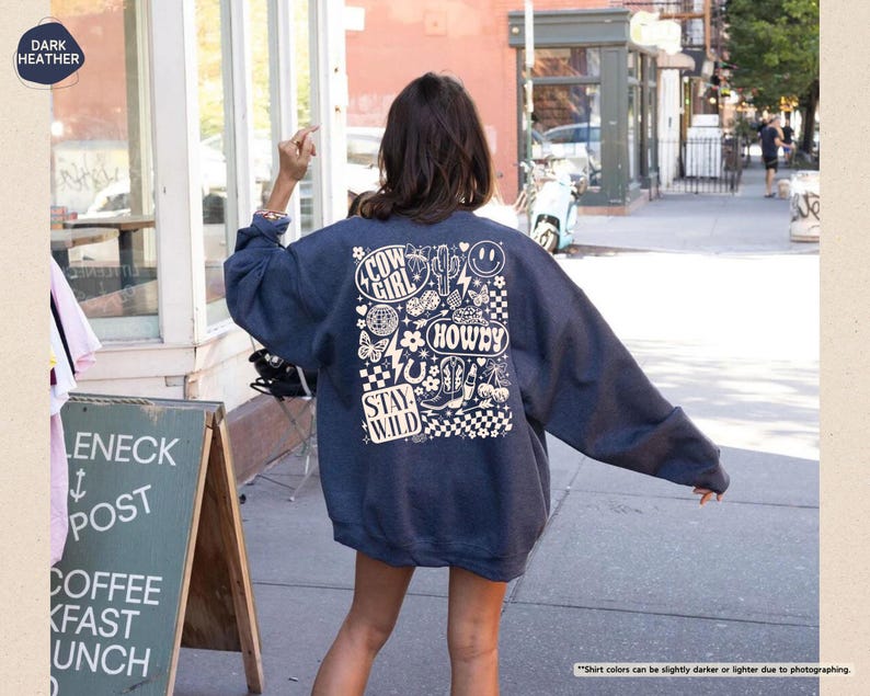 K&ouml;nnte beinhalten: Dunkelblau meliertes Sweatshirt mit wei&szlig;em Grafikdesign auf dem R&uuml;cken. Das Design enth&auml;lt die W&ouml;rter "Cow Girl", "Howdy" und "Stay Wild" sowie verschiedene Illustrationen zum Thema Western. Das Sweatshirt wird im Freien getragen.