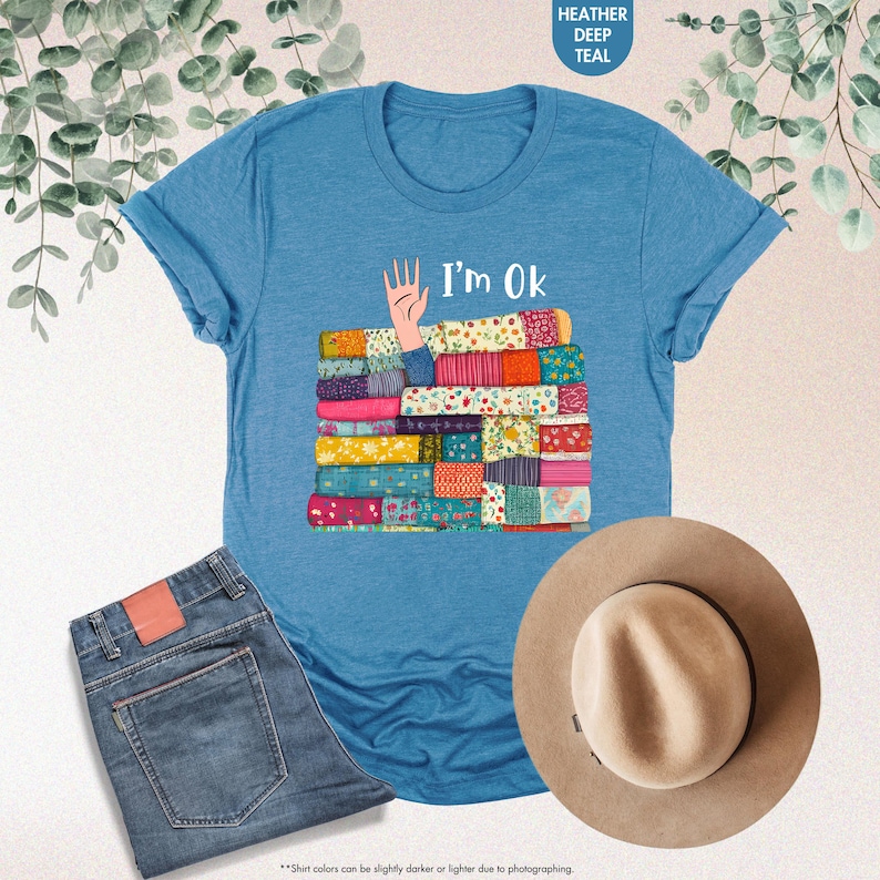 May include: Heather deep teal t-shirt with the text "I'm Ok" and a graphic of a hand and a stack of colorful fabric. The shirt is paired with blue jeans and a tan hat. The shirt's color may vary slightly.